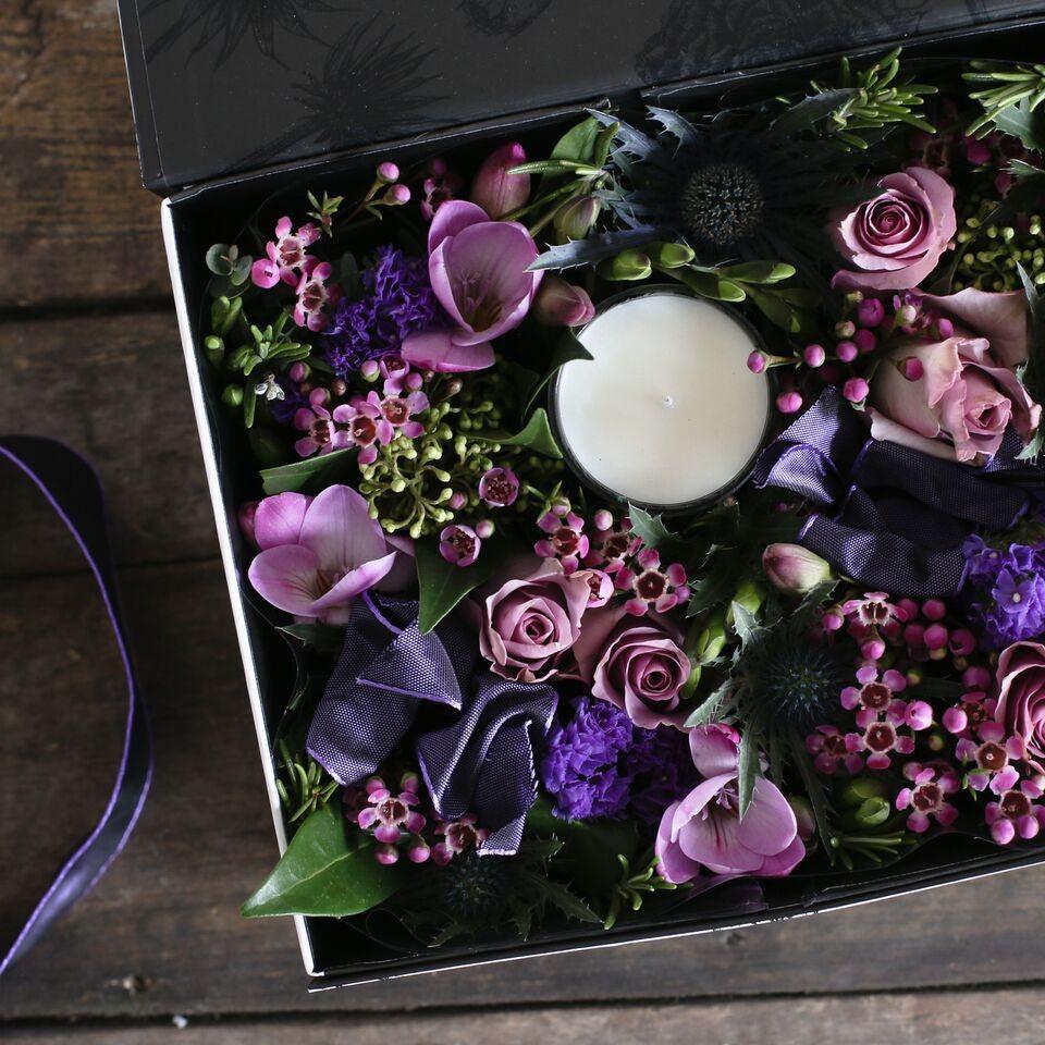 A gift box full of pink and purple flowers and a scented candle.