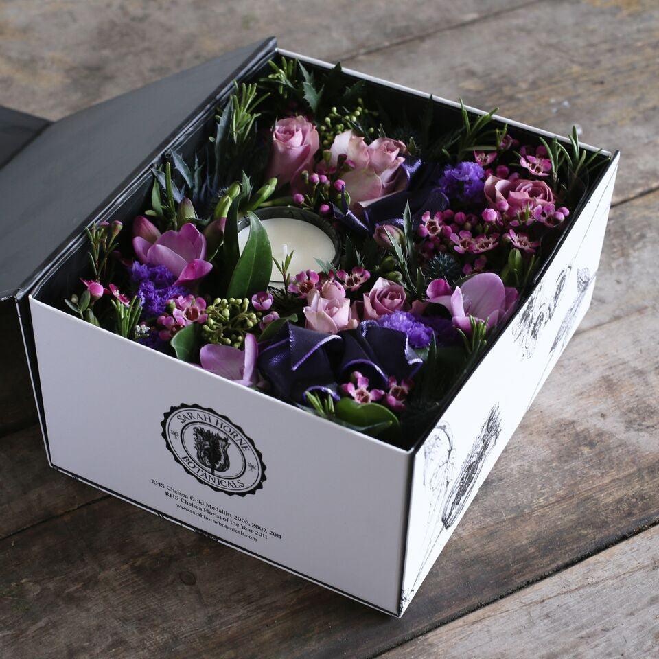 A gift box full of pink and purple flowers and a scented candle.