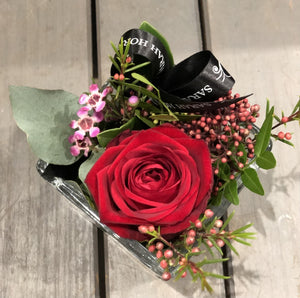 a single rose in a glass vase with foliage and ribbon.